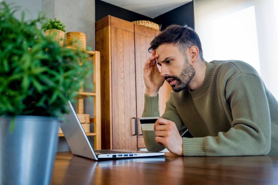 Worried man seated at a table holding a credit card while looking at a laptop screen, with one hand resting against the forehead. Wooden furniture, shelving, and houseplants are visible in the background