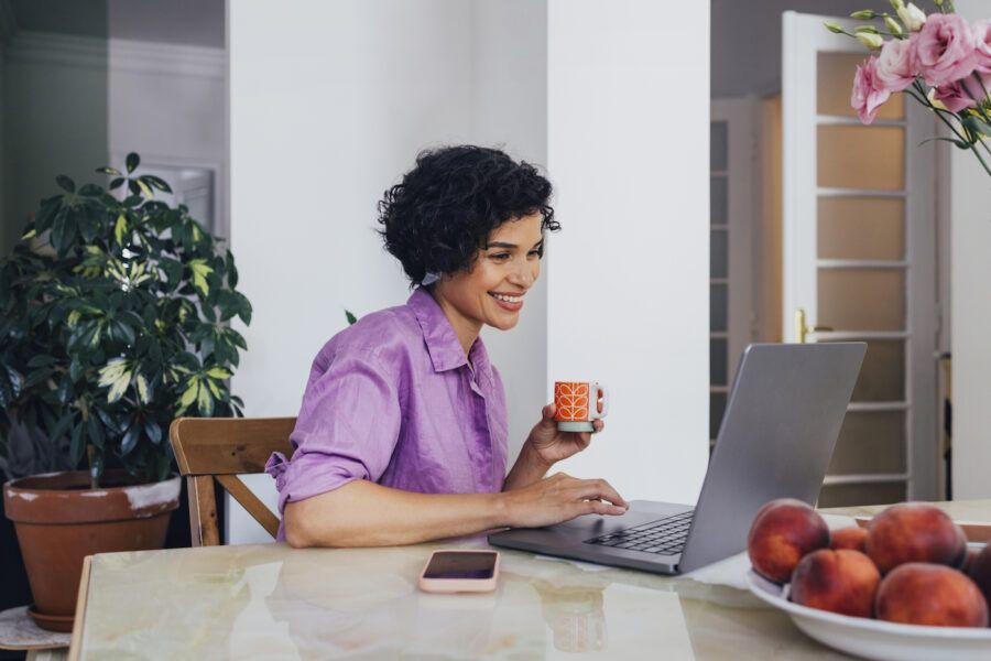 A woman smiling while scrolling on her laptop, drinking espresso from a colorful cup.