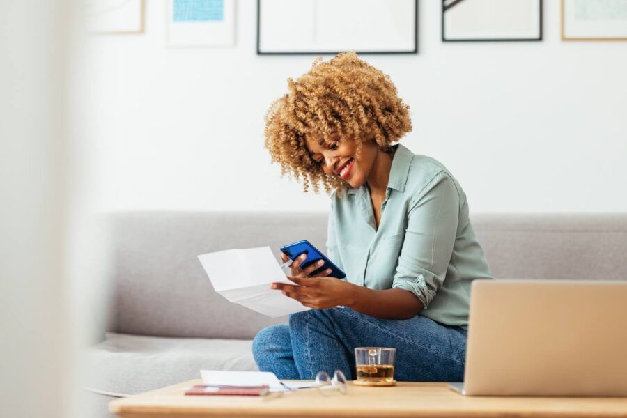 Smiling woman using her mobile phone to pay bills at home, with an open laptop and a glass on the coffee table beside her.