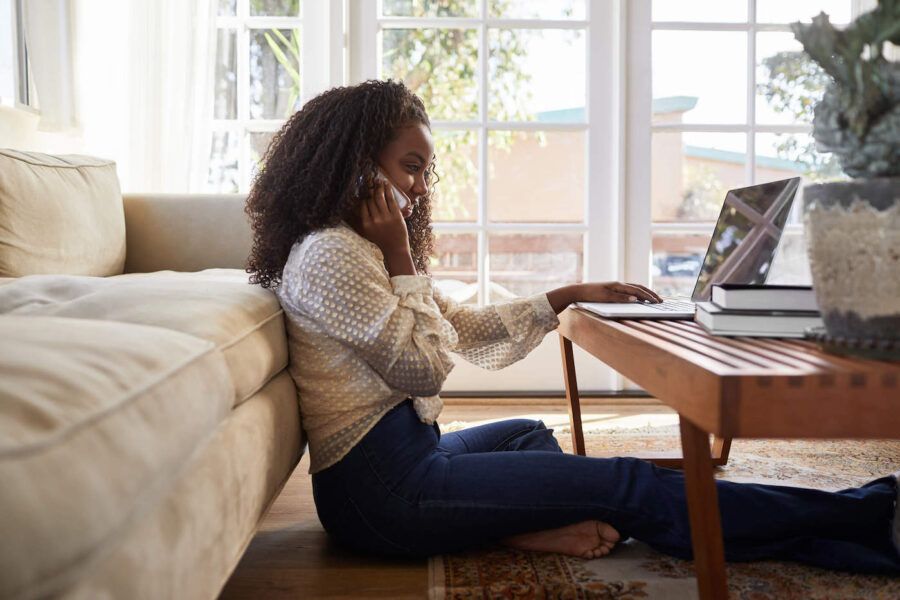 Young African American woman seated on the floor of her living room talking on the phone while browsing on laptop.