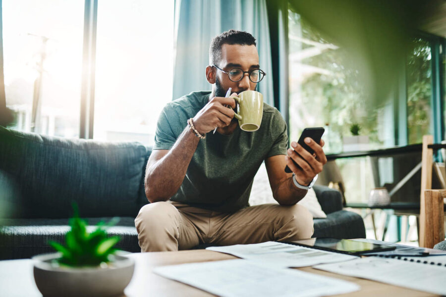 Young man sitting on couch looks at finances on phone while drinking from mug.