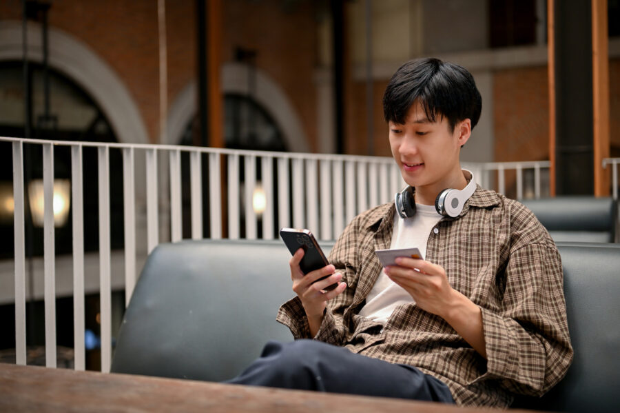 Happy young man sitting in outdoor booth table, smiling and holding smartphone in one hand and crebit card in the other.