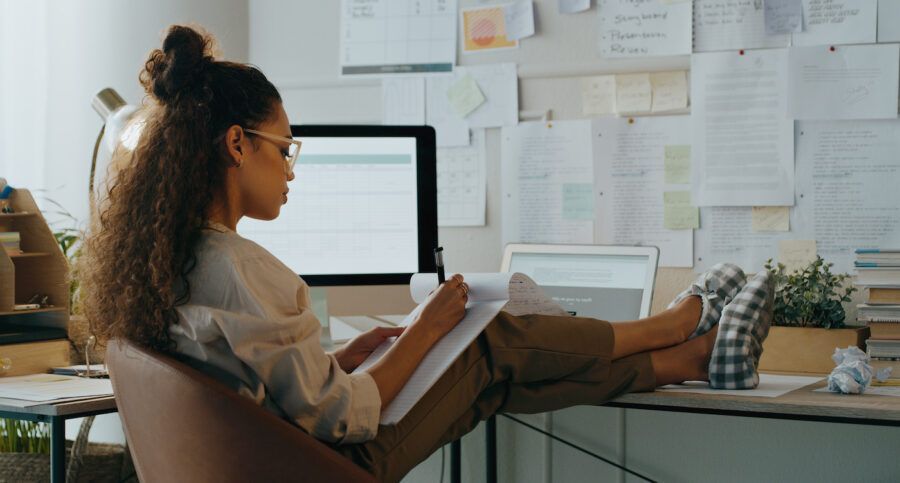 A young woman researching insurance in her office, writing on a notepad with her feet up on her desk.
