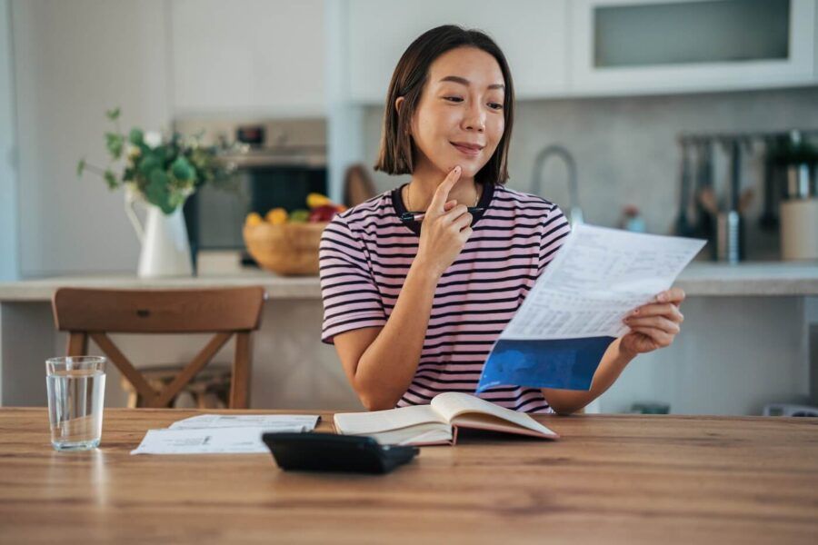 Woman sitting at a wooden table in a bright kitchen, holding and reading a printed document while resting one finger on the chin; a notebook, calculator, papers, and a glass of water are on the table, with kitchen items and shelves visible in the background.