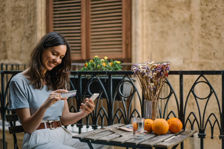 Young woman seated on a small table on a balcony looking at her credit card while holding a phone.