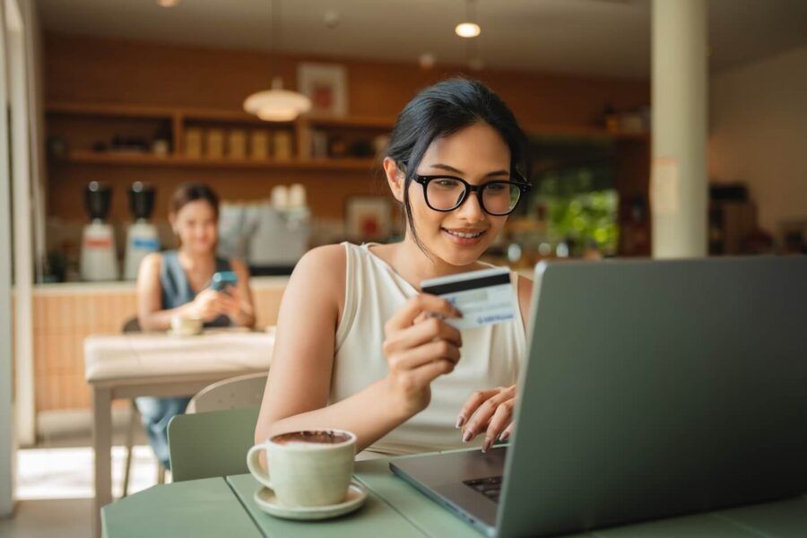 Smiling young woman seated at a café table holding a credit card while using a laptop. A cup of coffee sits on the table, and another person is visible in the background using a smartphone. Warm lighting, coffee shop décor, and café equipment are visible in the background