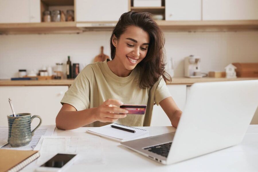 Smiling young woman seated at a kitchen table holding a credit card while using a laptop, with a notebook, smartphone, coffee mug, and paperwork nearby. Kitchen cabinets and countertops are visible in the background.