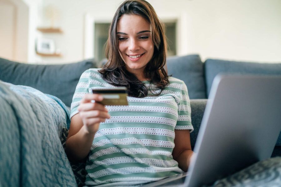 Smiling young woman sitting on a sofa at home using a laptop while holding a credit card, making an online purchase in a comfortable living room setting.