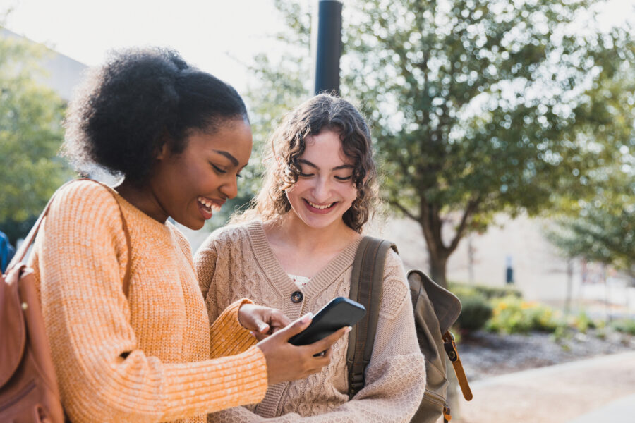 A teenage girl shows her female friend something on her smart phone that makes both of them smile.